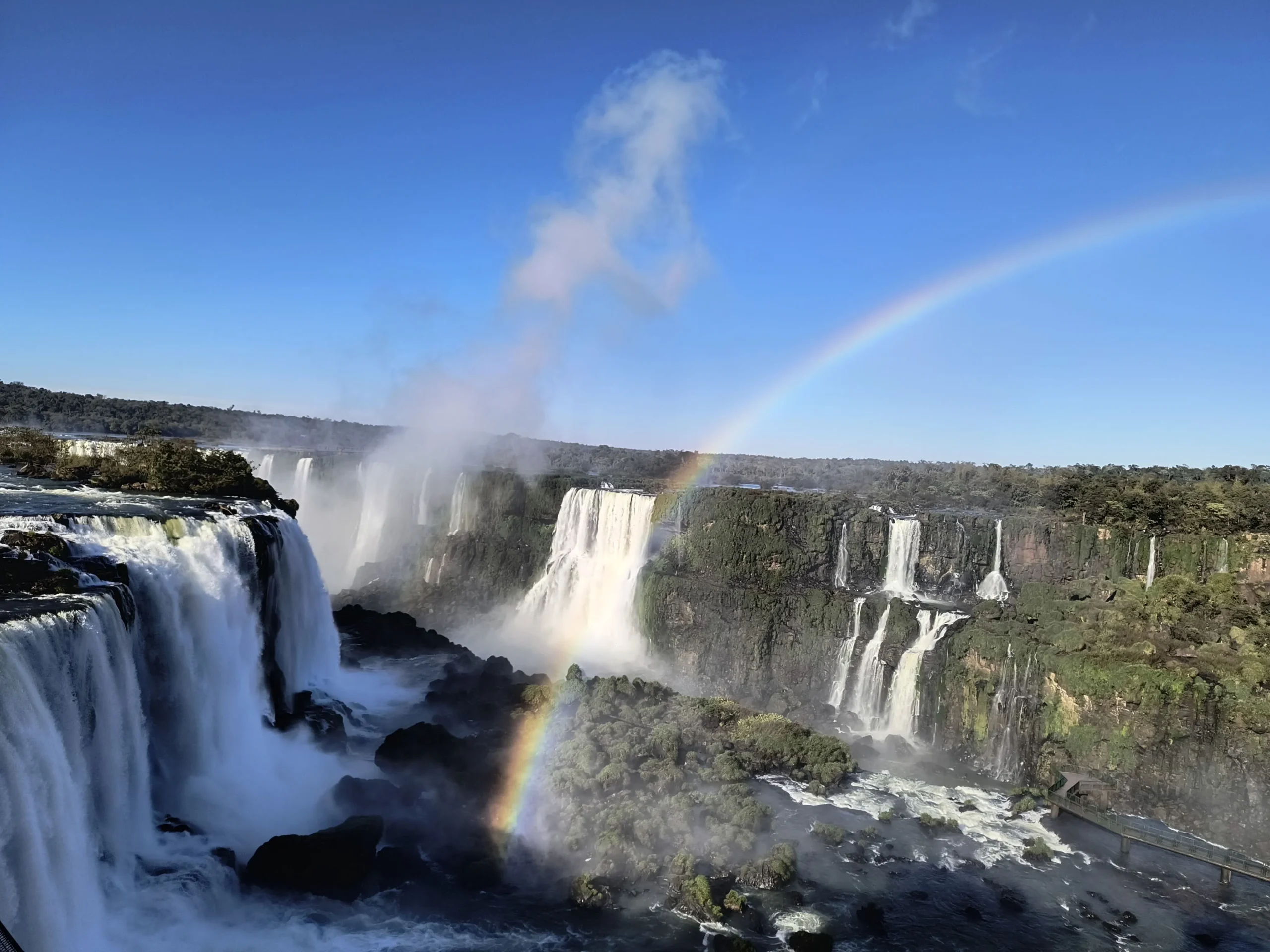 The magnificent Iguazu Falls from the Brazilian perspective, adorned with a vibrant rainbow, a highlight of Miguel's tours.